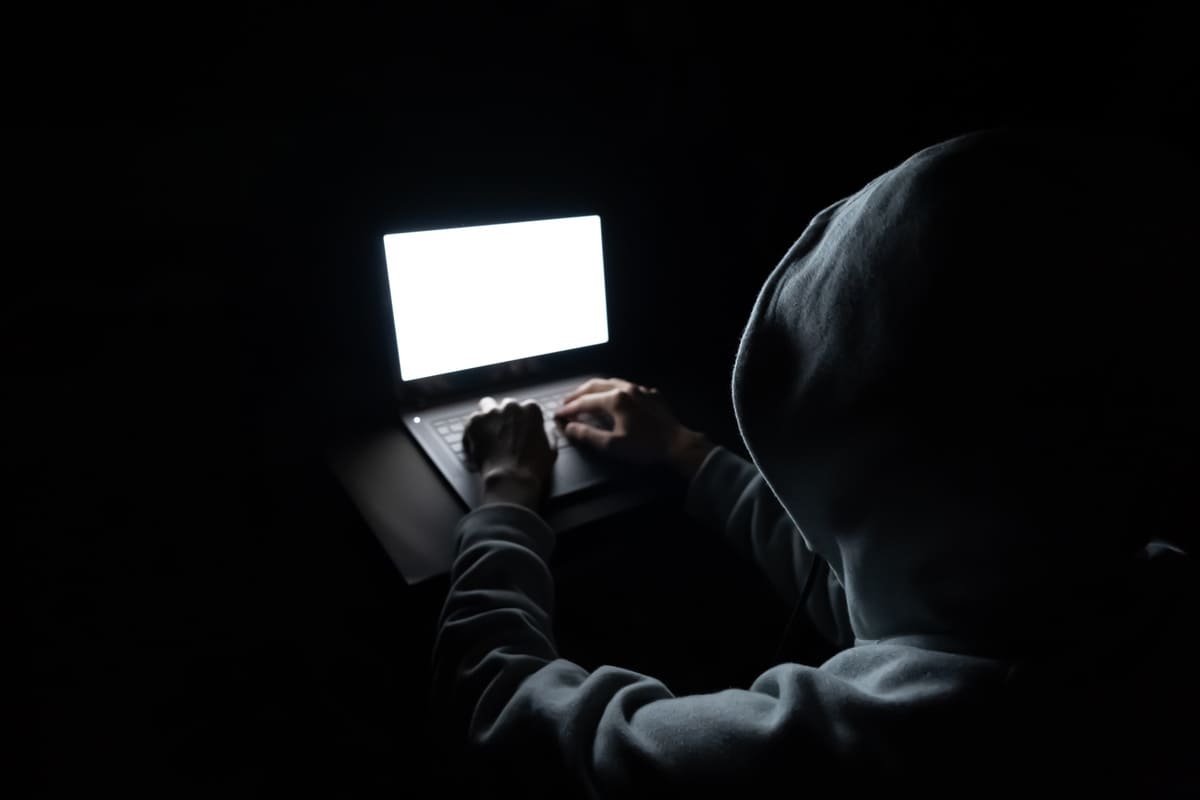 Hooded figure at a laptop in the dark representing cyber fraud