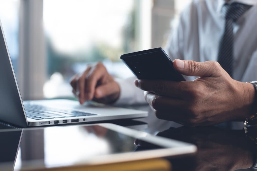 A person in a white dress shirt and tie sits at a desk holding a smartphone.