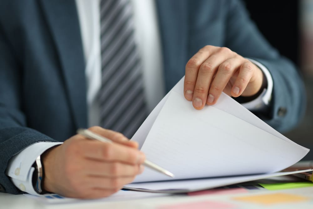 A person wearing a suit, white shirt, and striped tie is seated at a desk, using one hand to lift a page of a document while holding a pen in the other hand.