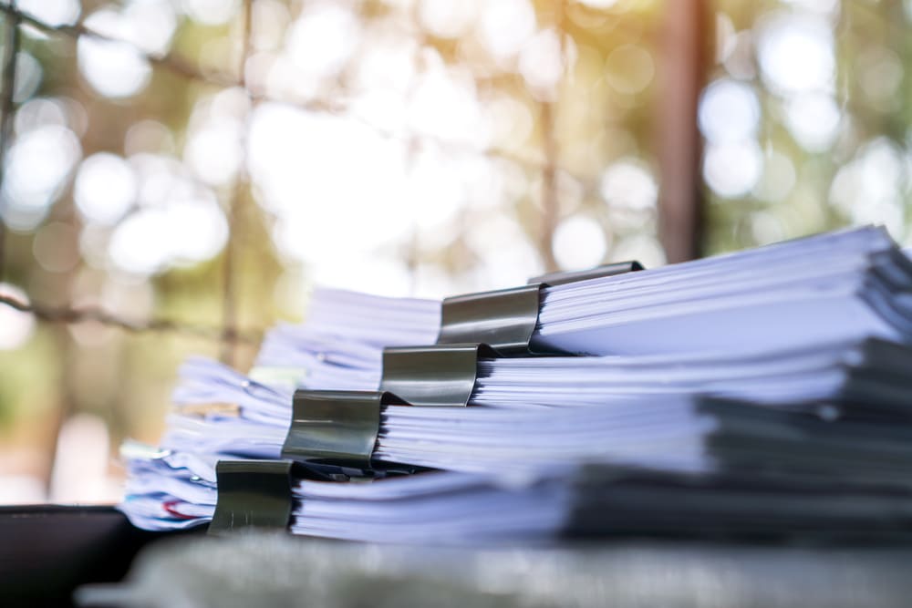 A close-up shot shows multiple stacks of white papers secured with black binder clips, sitting on a surface with a brightly lit, out-of-focus background.