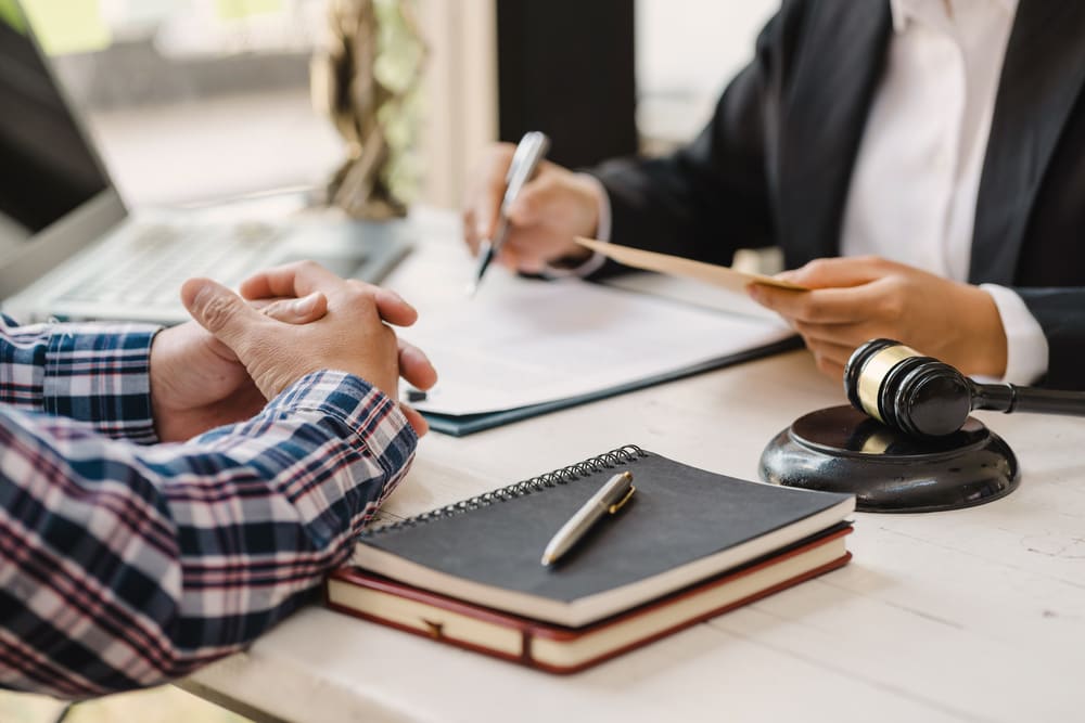 A person wearing a plaid shirt sits with clasped hands across a desk from another individual in a suit who is writing on a document.