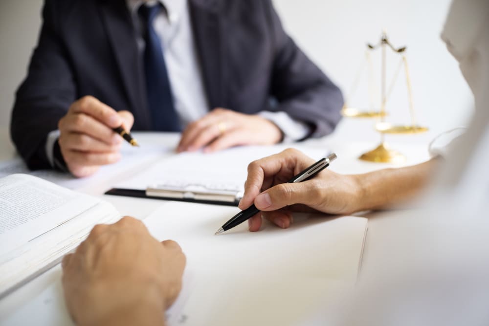 A person in the foreground writes on a document with a pen at a desk, while another person in a suit and tie sits across from them, also holding a pen.