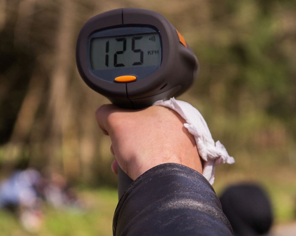 A close-up, first-person perspective shot shows a person's hand holding a handheld radar speed gun