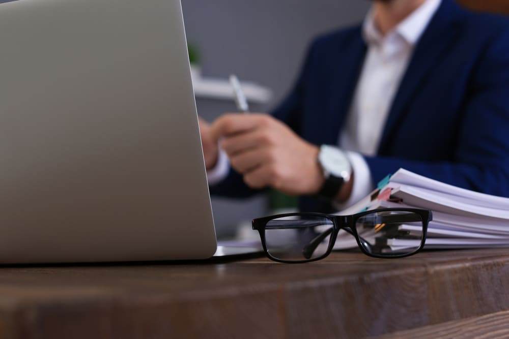 A pair of black-rimmed glasses rests on a wooden desk in the foreground, next to a laptop computer and a stack of papers