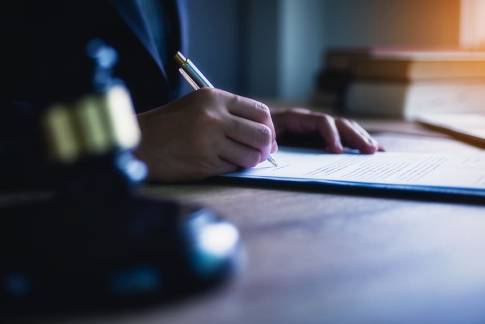 a person seated at a wooden desk is writing on a document with a pen.